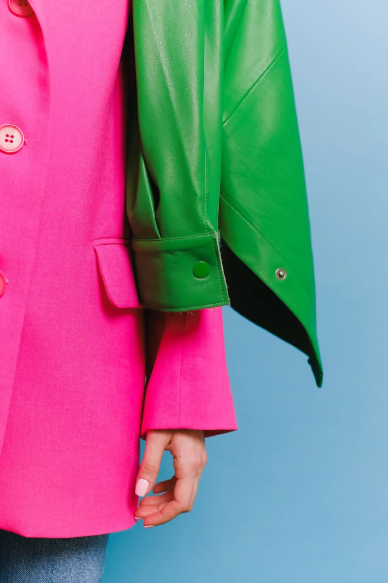 Detail of a bright pink blazer layered with a green leather jacket against a pale blue background.