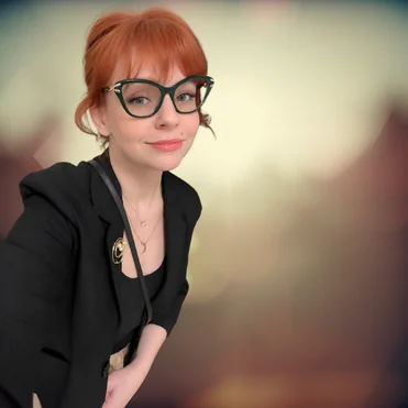 Audrey Mast with red hair and green glasses, wearing a black jacket against a softly blurred background.