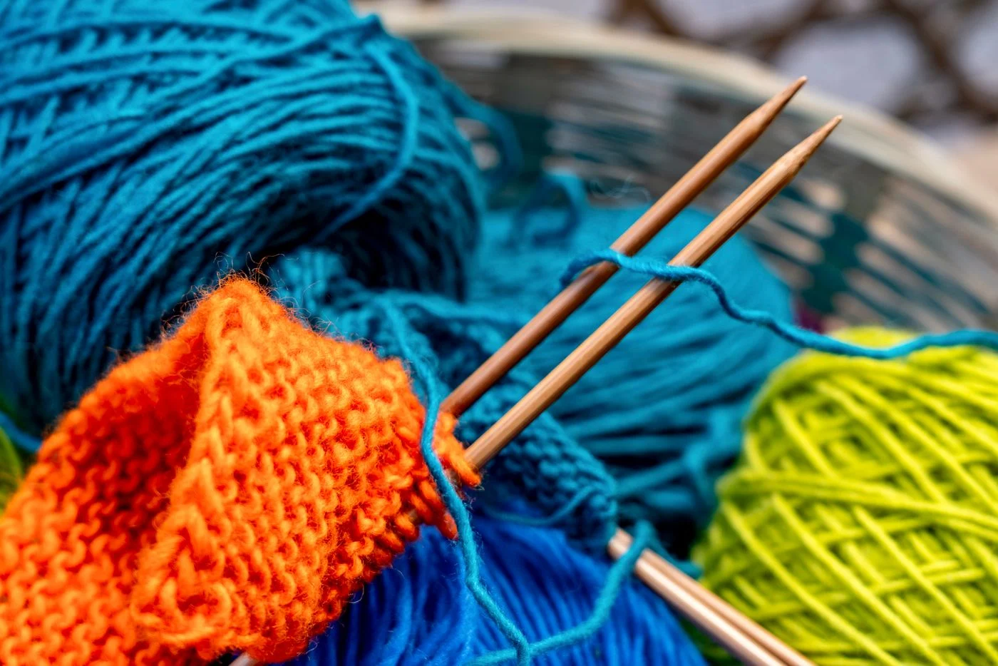 Orange knitting on needles beside turquoise and yellow yarn.