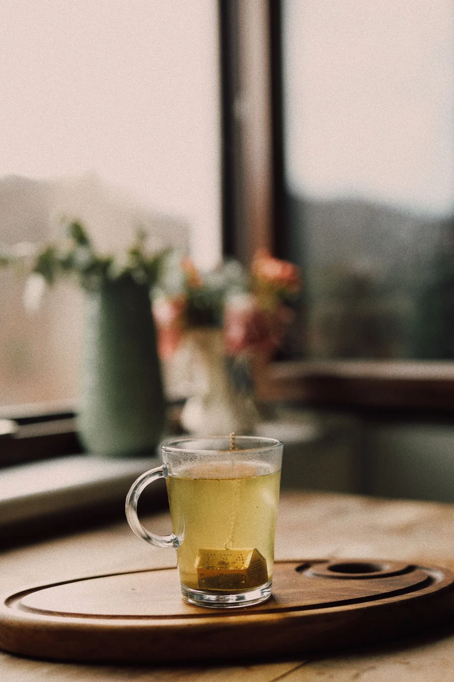 A glass cup of tea on a wooden tray beside a window.
