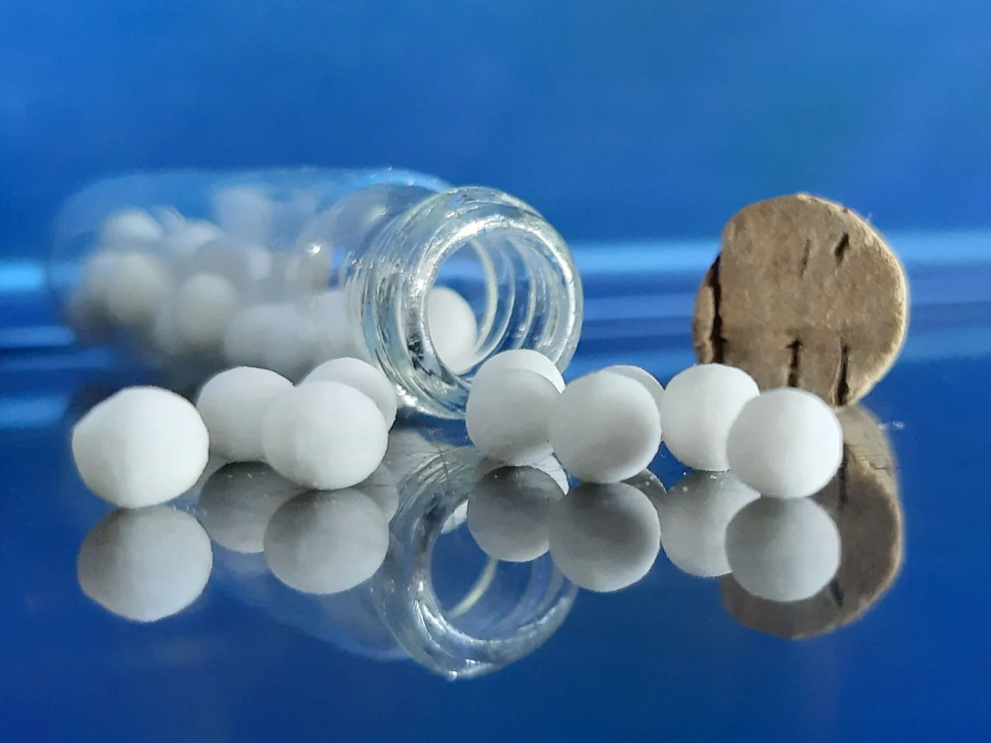 White homeopathic globules spilling from a glass bottle onto a reflective blue surface.