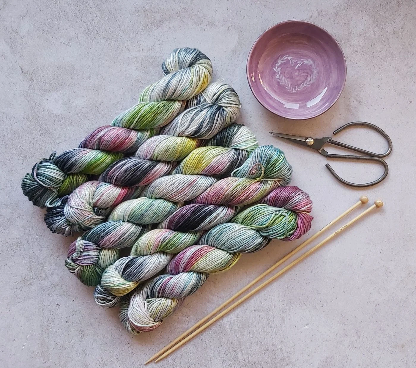 Pastel skeins of yarn beside knitting needles, scissors, and a pink bowl.